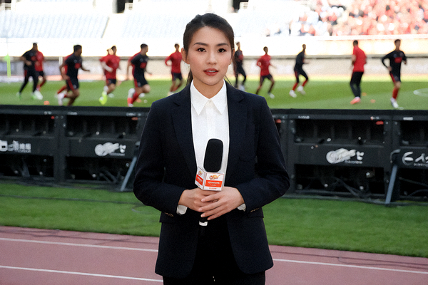 体育新闻记者在球场边进行赛前报道，背景是正在热身的球员